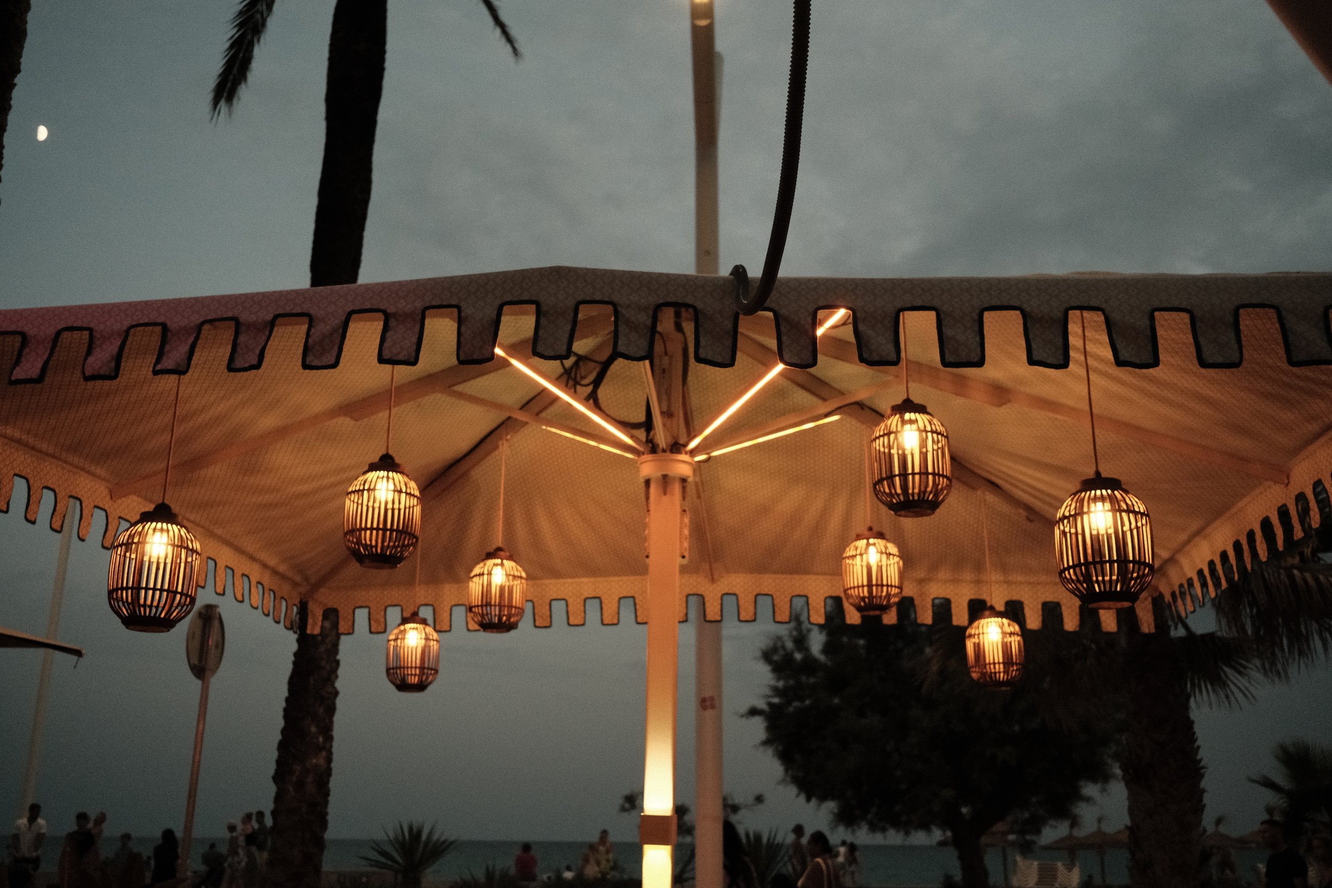 Lit lanterns hanging from a beach parasol at dusk